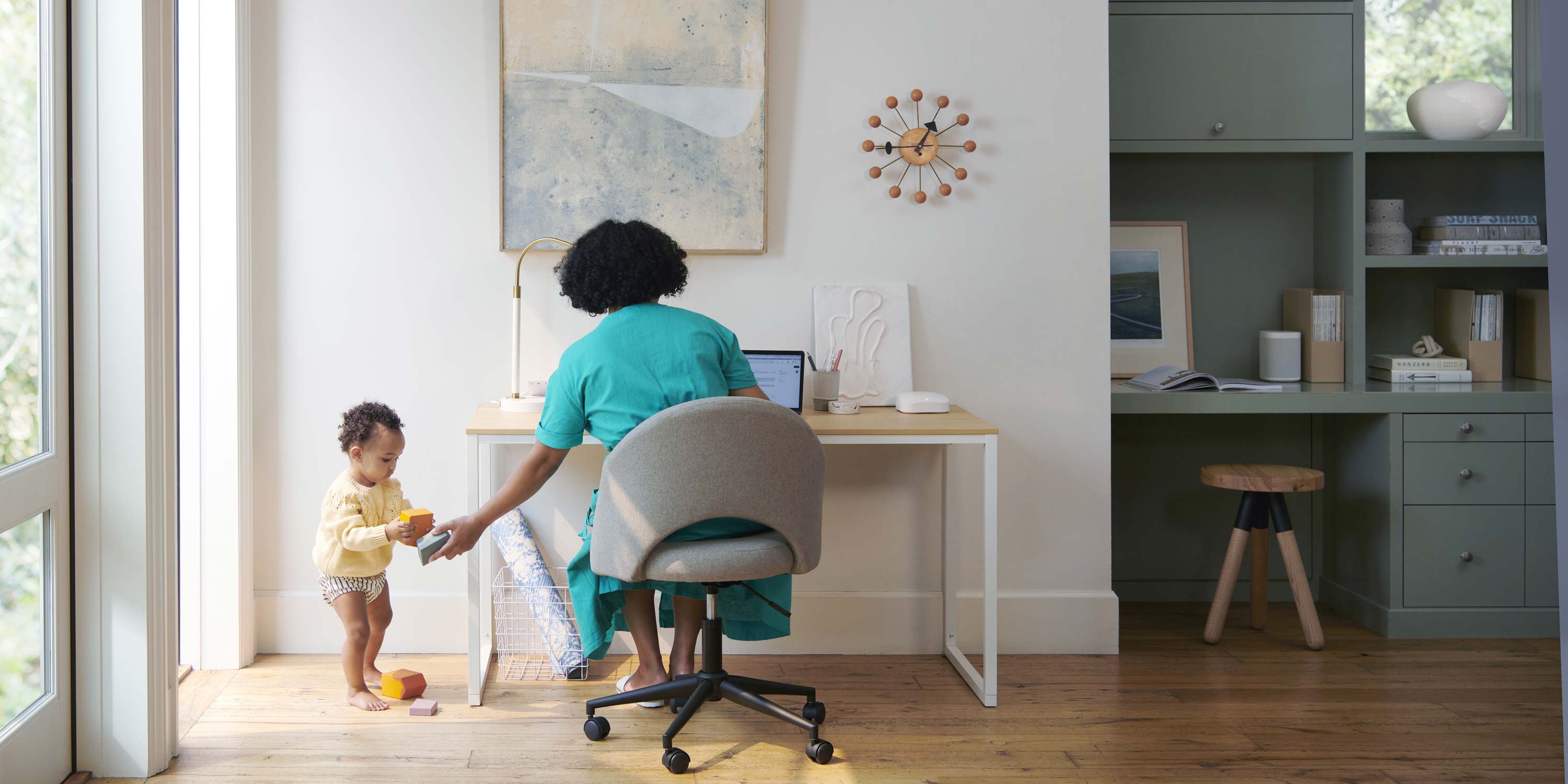 woman in a teal dress playing with a toddler while she works in a nice home office