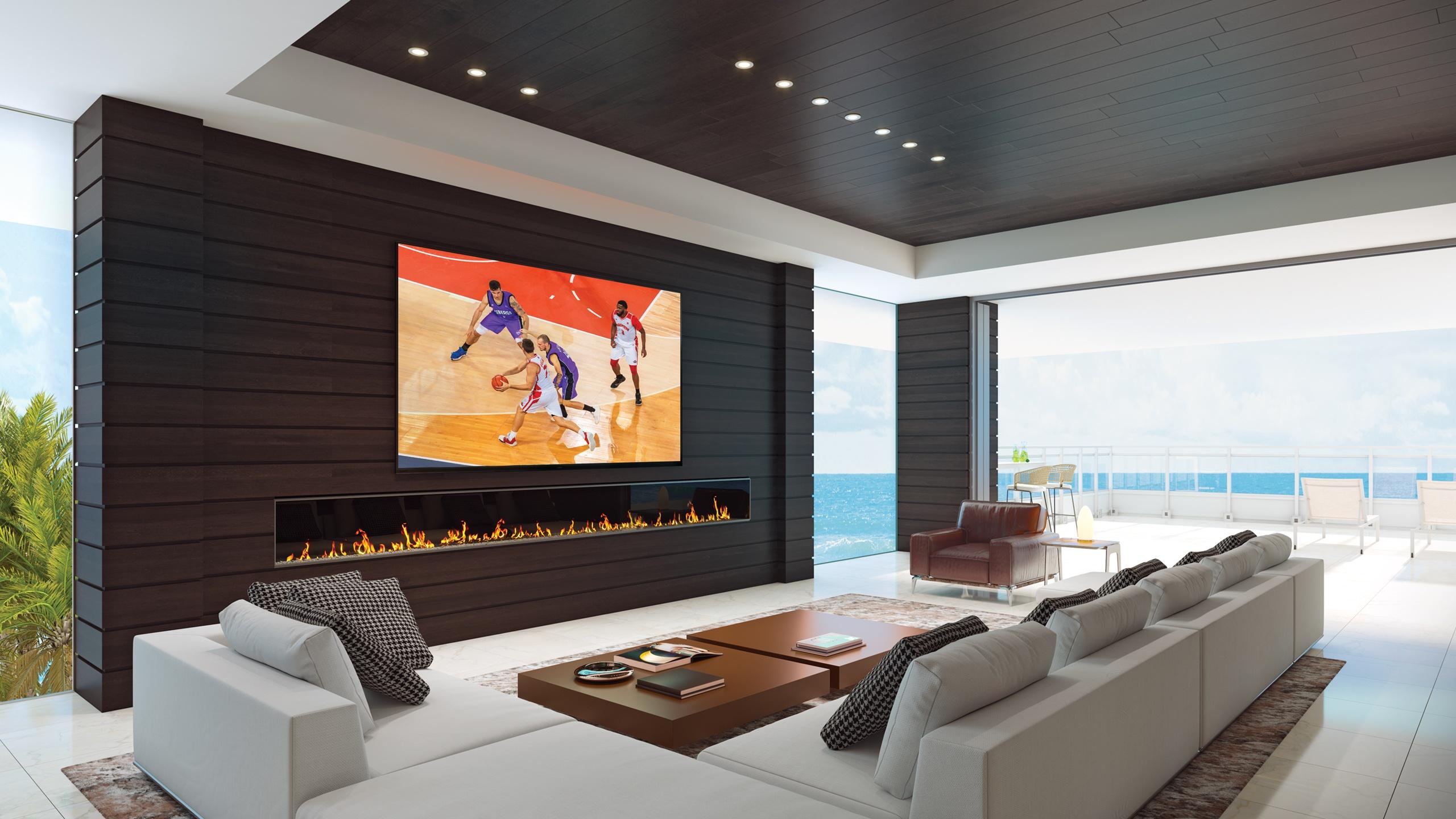 A luxurious living room featuring a large Sony TV on a dark wood accent wall, a modern fireplace below, and a view of the ocean.