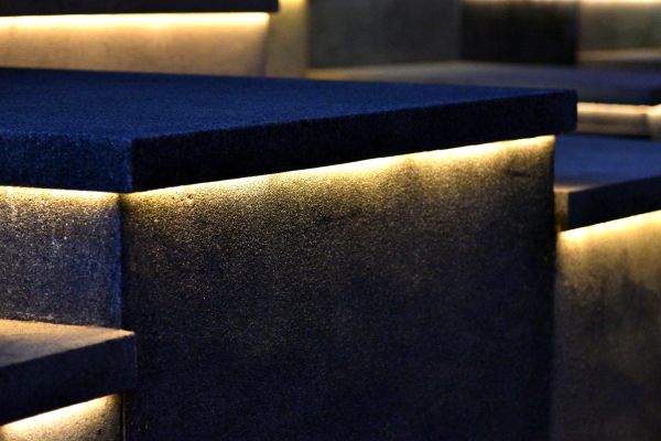 A modern architectural building illuminated at night with strategically placed outdoor lighting, highlighting the stone textures, wooden elements, and structural details against a mountainous backdrop. The warm lighting creates a welcoming ambiance while emphasizing the building's design features.