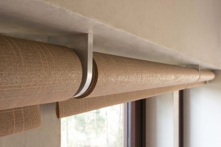 A close-up view of a beige woven roller shade mounted on a metal bracket, installed above a window with a light-textured wall and a glimpse of greenery outside.