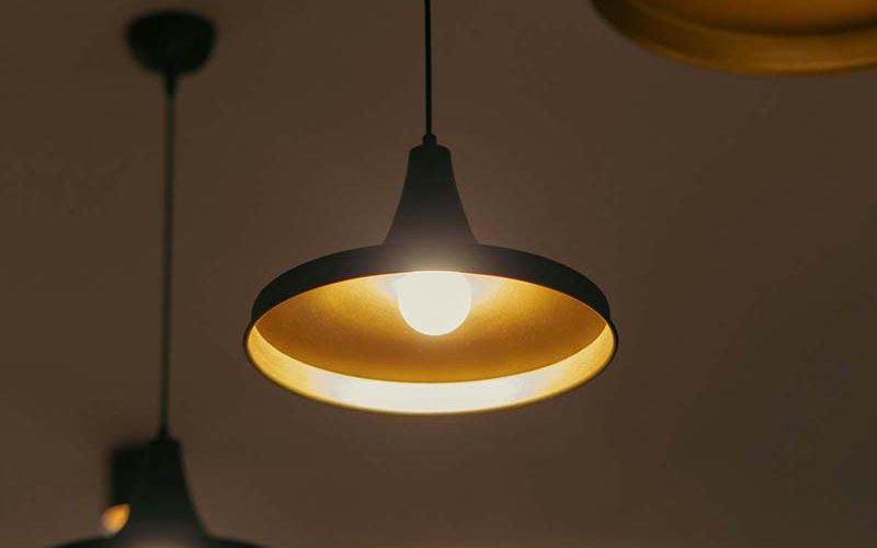 A cozy and elegant living space featuring soft ambient lighting with modern pendant lights, recessed ceiling fixtures, and a unique sculptural light over the kitchen. The room includes plush seating, round tables, and neutral tones, creating a warm and inviting atmosphere.